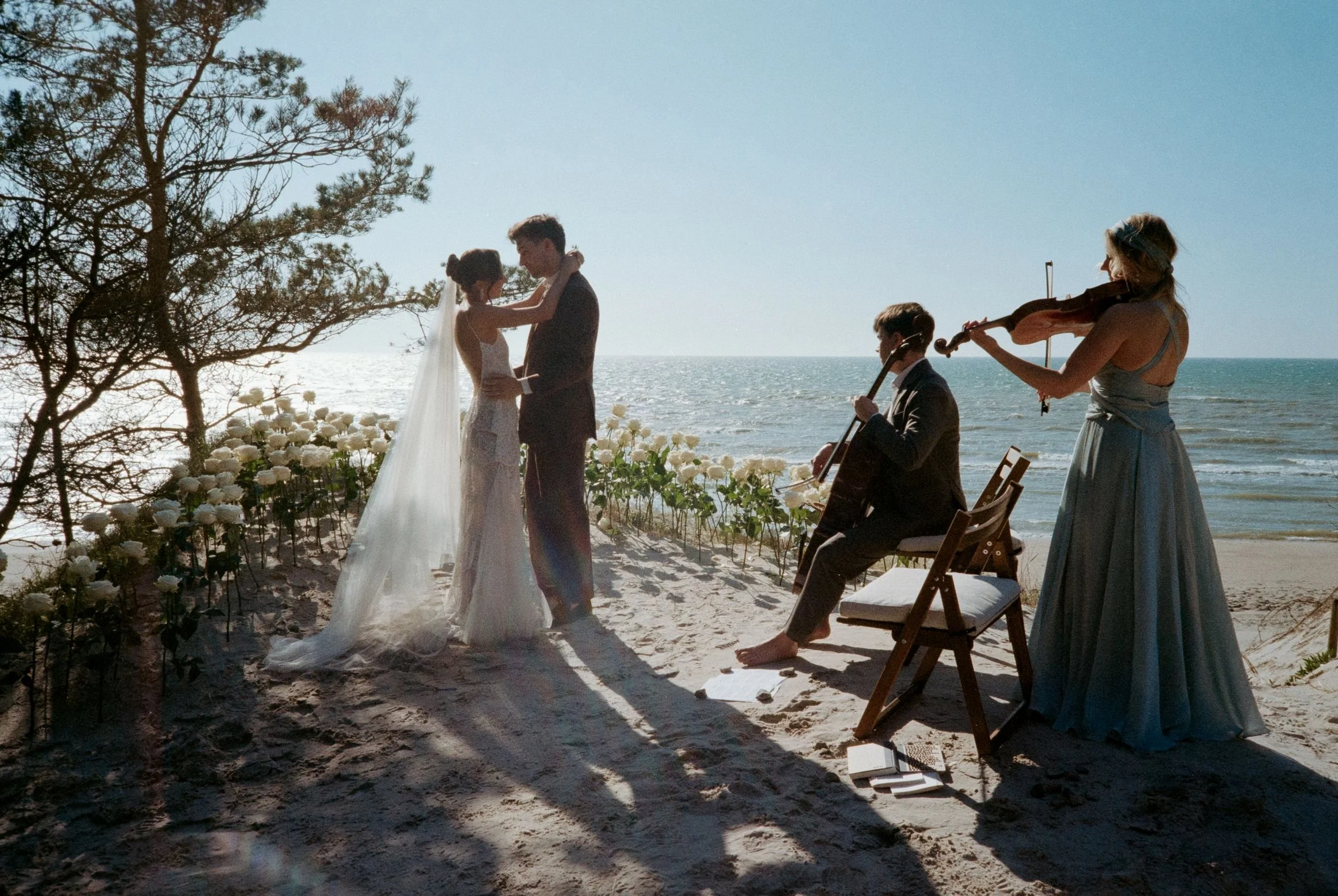 A couple getting married on a beach with a musician playing the violin and another playing the cello nearby, during sunset over the ocean.