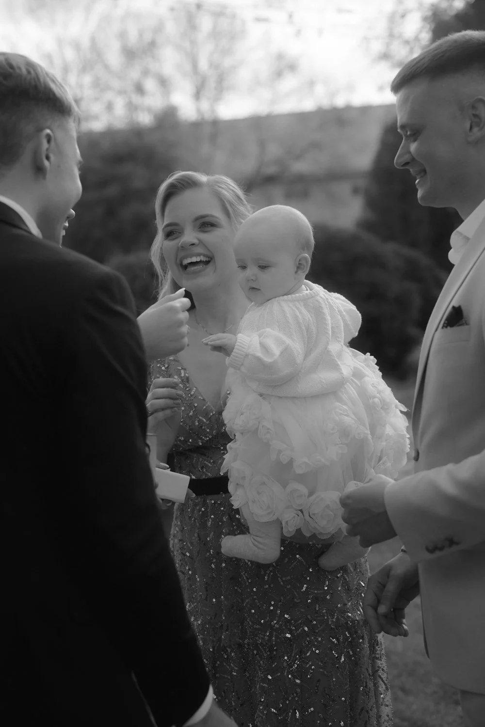 A black and white photo of a family gathered outdoors, with a woman holding a young child dressed in a frilly dress. Two men, dressed in suits, are nearby; one is smiling at the group, and the other appears to be speaking to them.