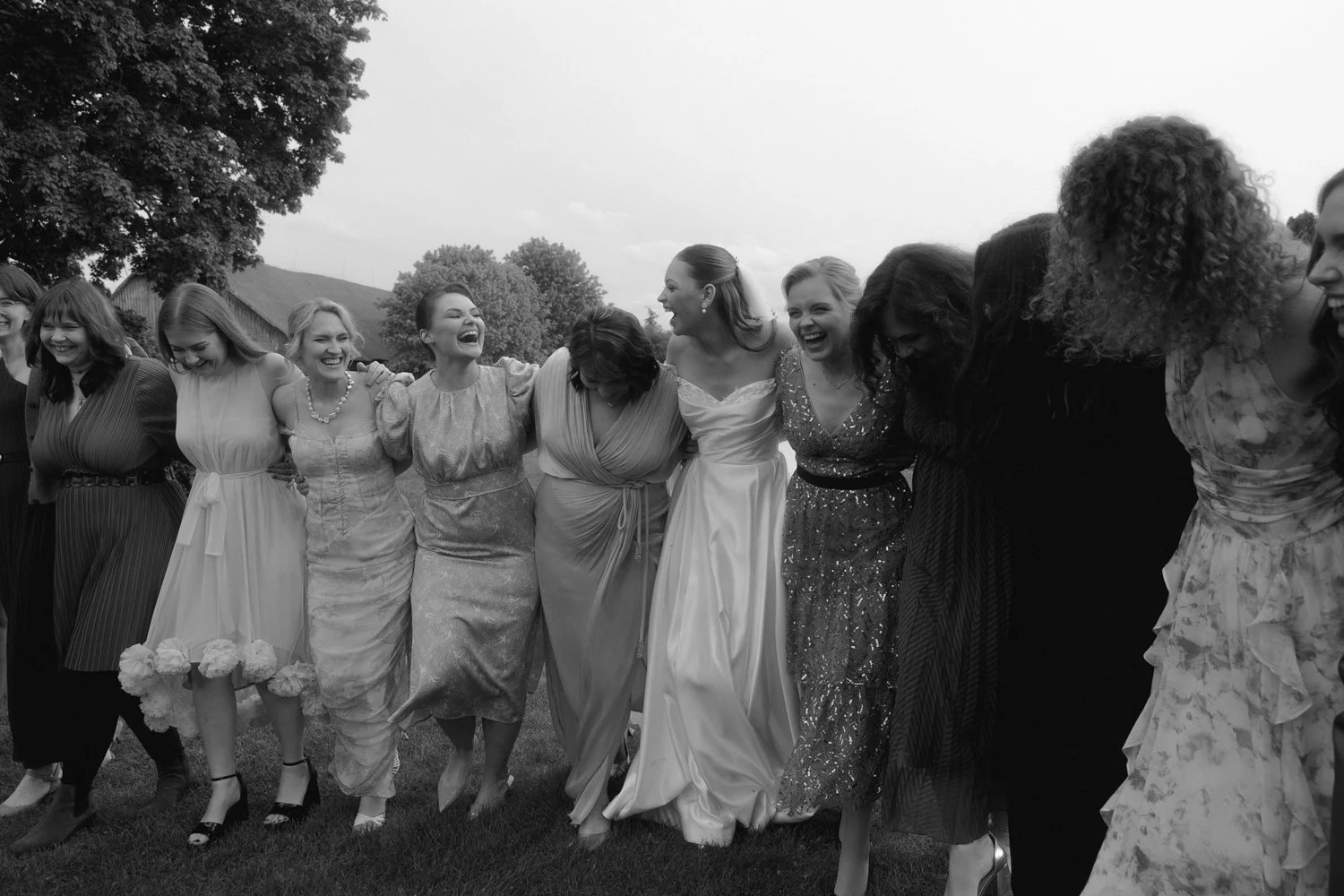 Group of women in dresses, smiling and laughing, standing outdoors on grass with trees and sky in background.