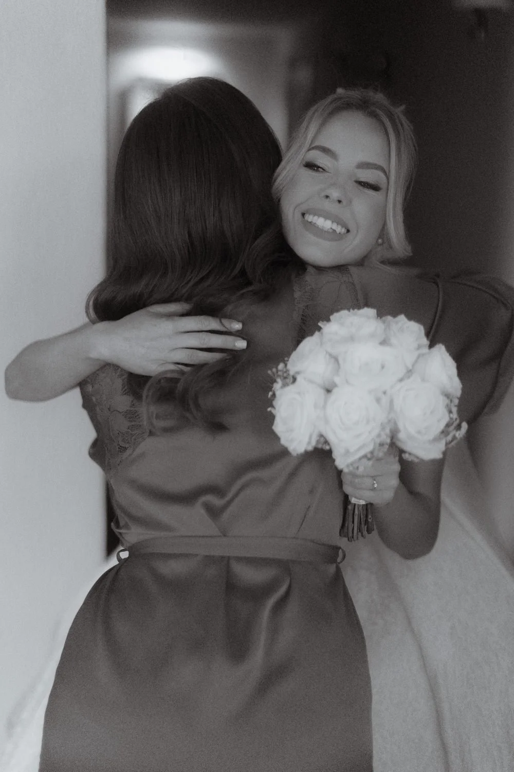 Two women hugging, one holding a bouquet of white roses. The woman facing the camera is smiling and has short, wavy hair. The other woman's face is not visible.