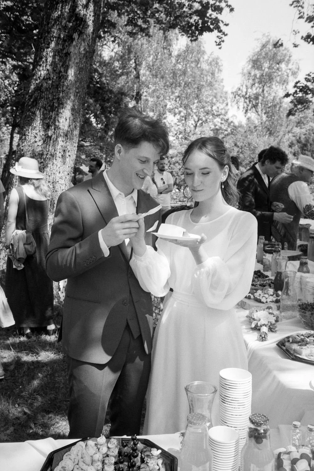 A black-and-white photo of a young man in a suit and a young woman in a white dress at an outdoor gathering, examining a dessert plate. There is a table with food and drinks in front of them, and other people are socializing in the background under t