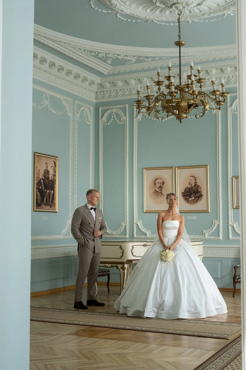A bride in a white wedding gown holding a bouquet stands next to a groom in a gray suit inside an elegant, ornately decorated room with framed portraits on the walls and a chandelier hanging from the ceiling.