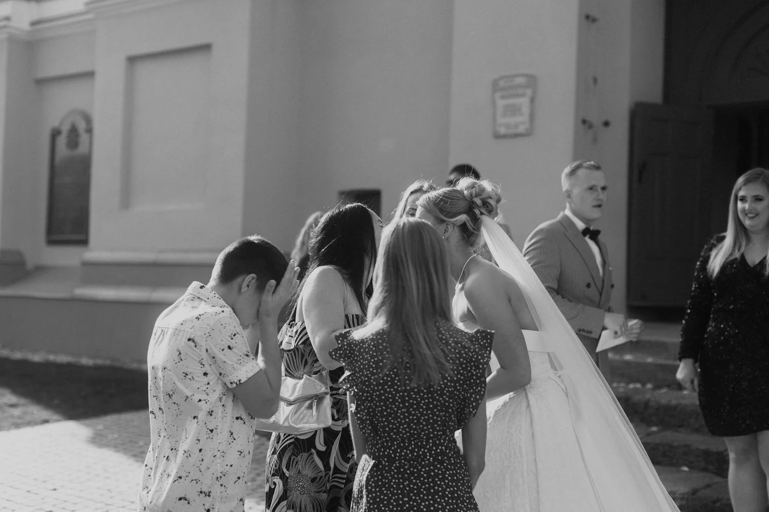 Black and white photo of a bride in a wedding dress talking to a group of women, while a boy covers his face with his hands nearby. The scene appears to be outside a church.