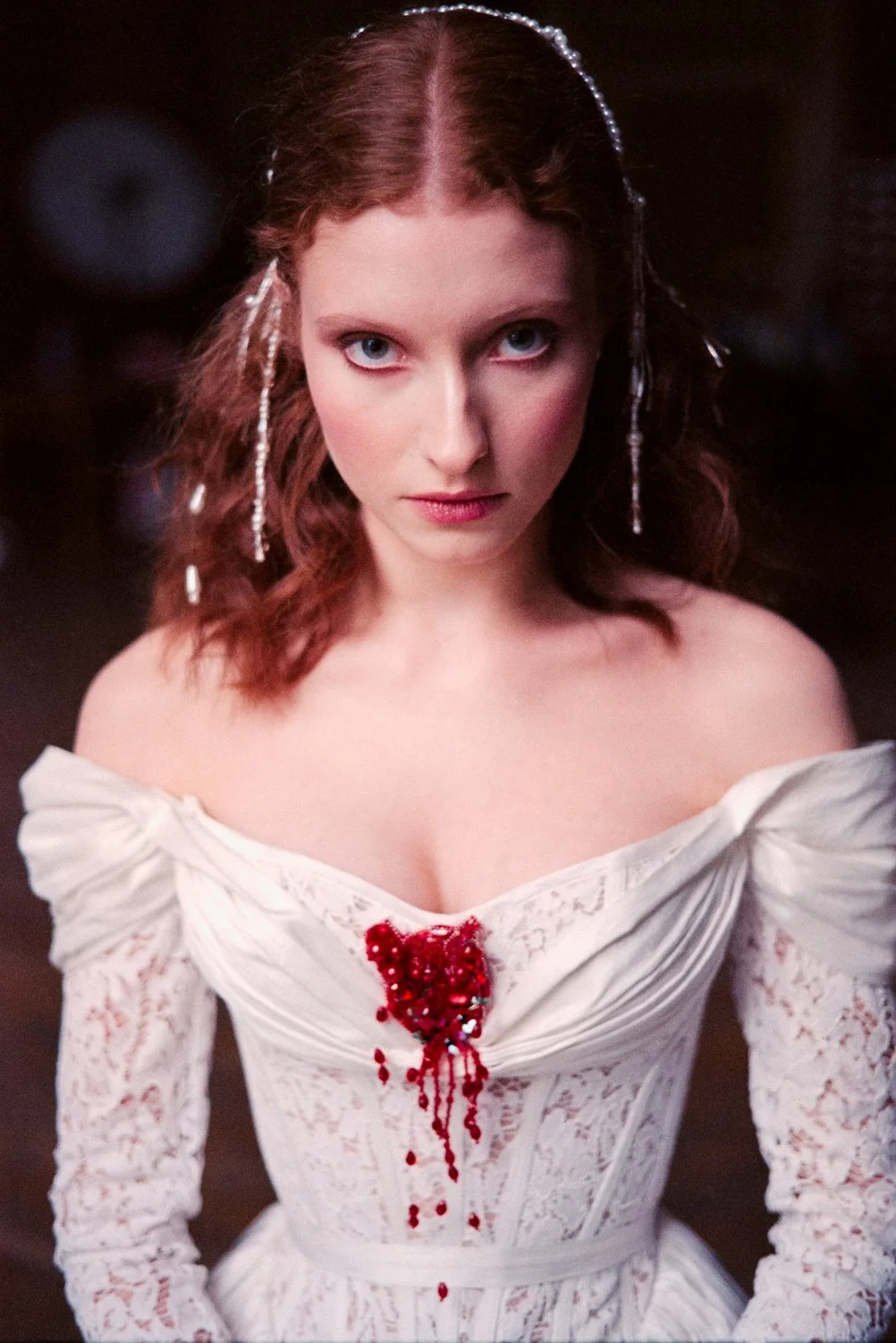A woman with red hair and blue eyes wearing a white off-shoulder dress with red blood-like embellishments on the chest, looking directly at the camera.