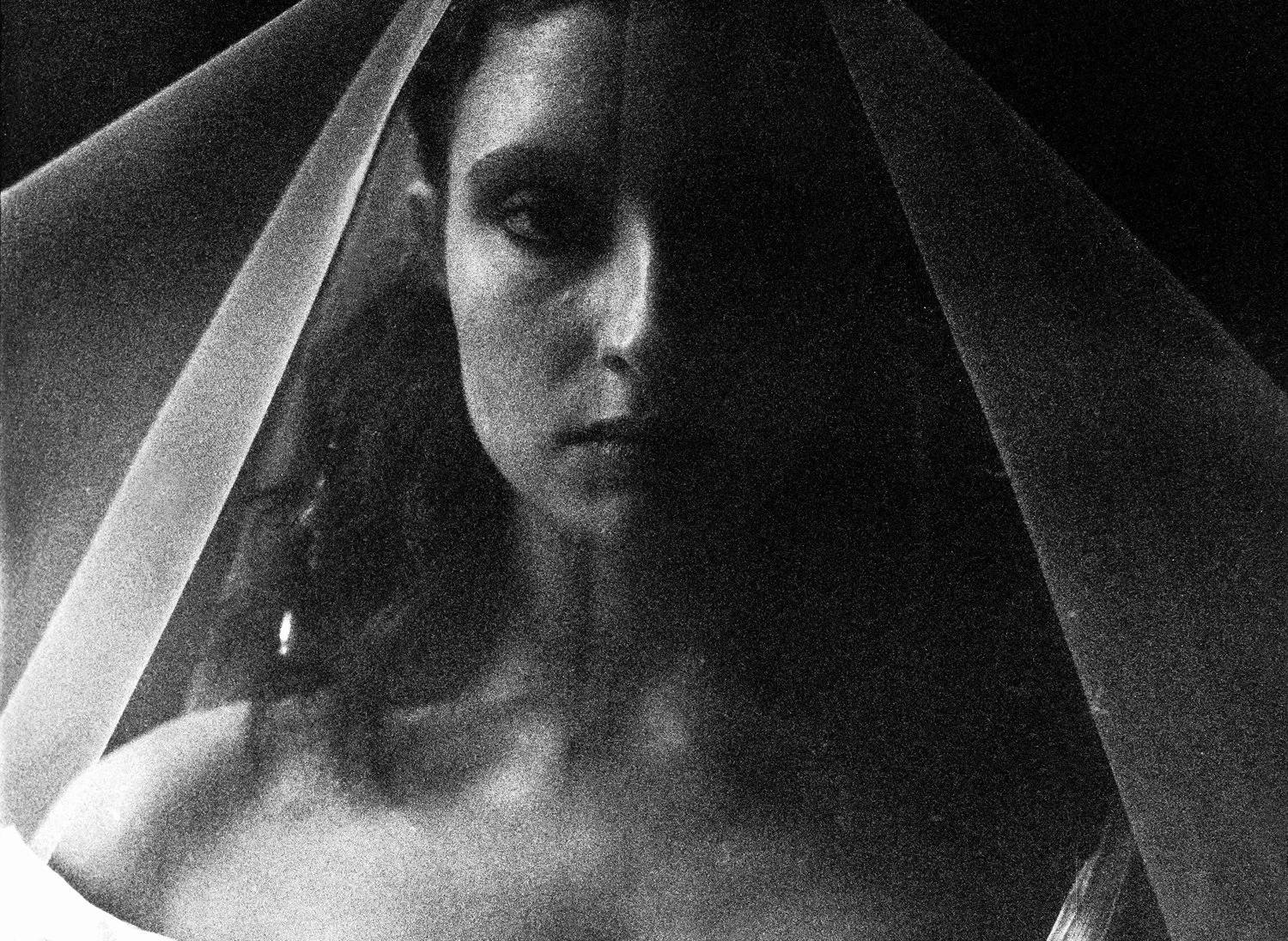 Black and white photo of a woman with dark hair, partially obscured by a sheer fabric, with one eye visible and a serious expression.