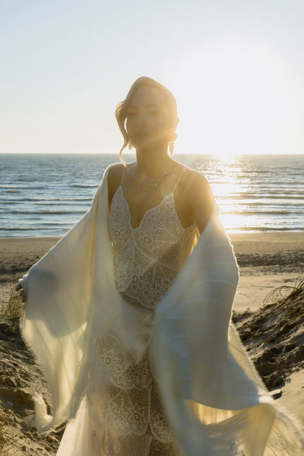 A woman in a patterned dress and earrings stands on a beach during sunset, with the ocean and the setting sun in the background.