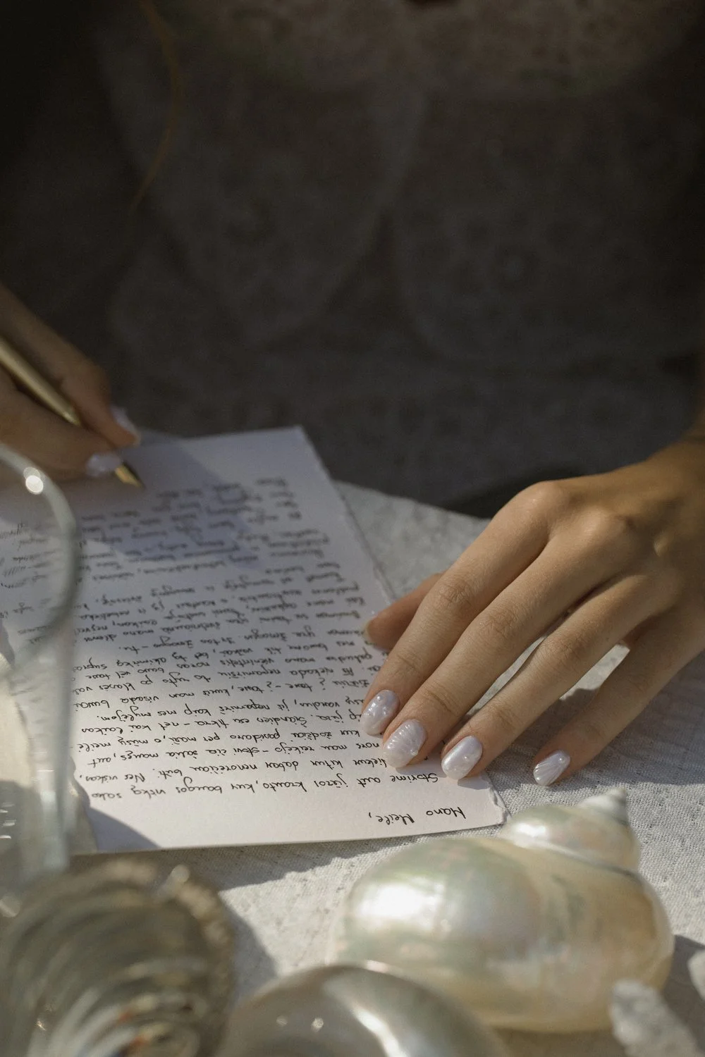 A person with light-colored nails is reading a handwritten letter on a table, with seashells and a misting spray bottle nearby.