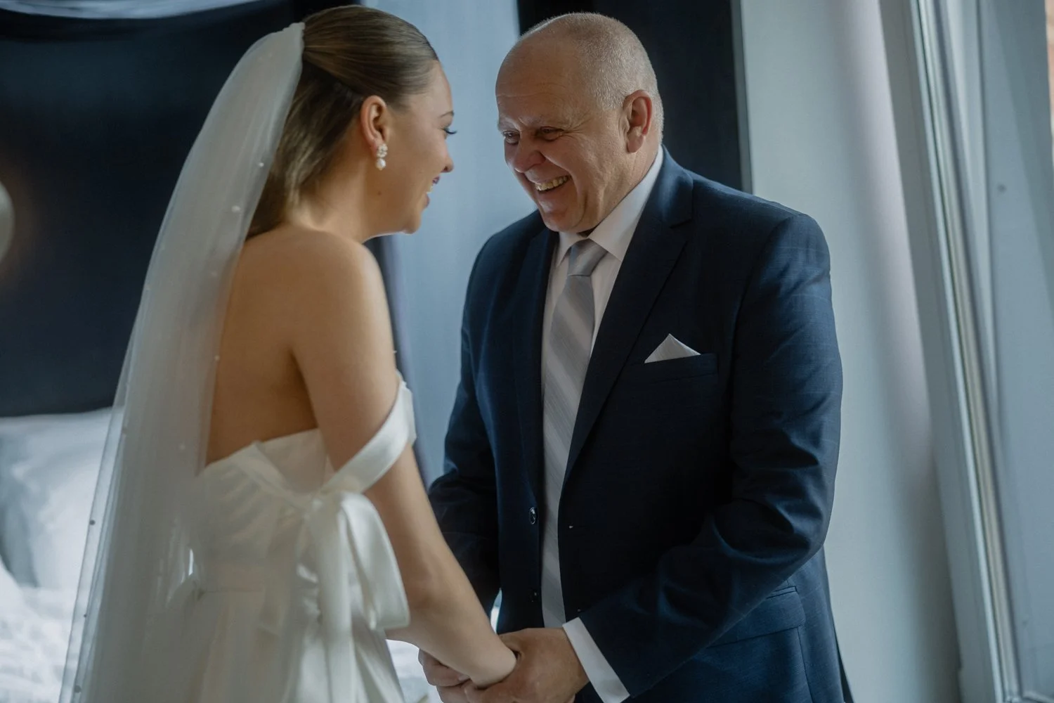 A bride and an older man, possibly her father, holding hands and smiling at each other in a bright room near a window.