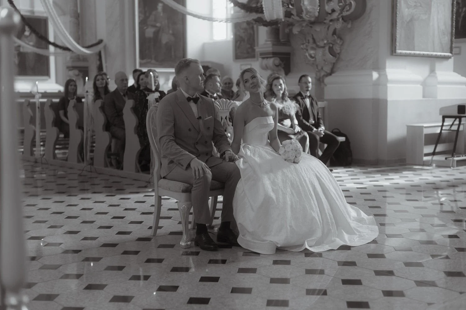 A black and white photograph capturing a wedding ceremony. The bride and groom are seated in the front, holding hands, with the bride holding a bouquet of flowers. The bride is wearing a strapless wedding gown, and the groom is dressed in a light-col