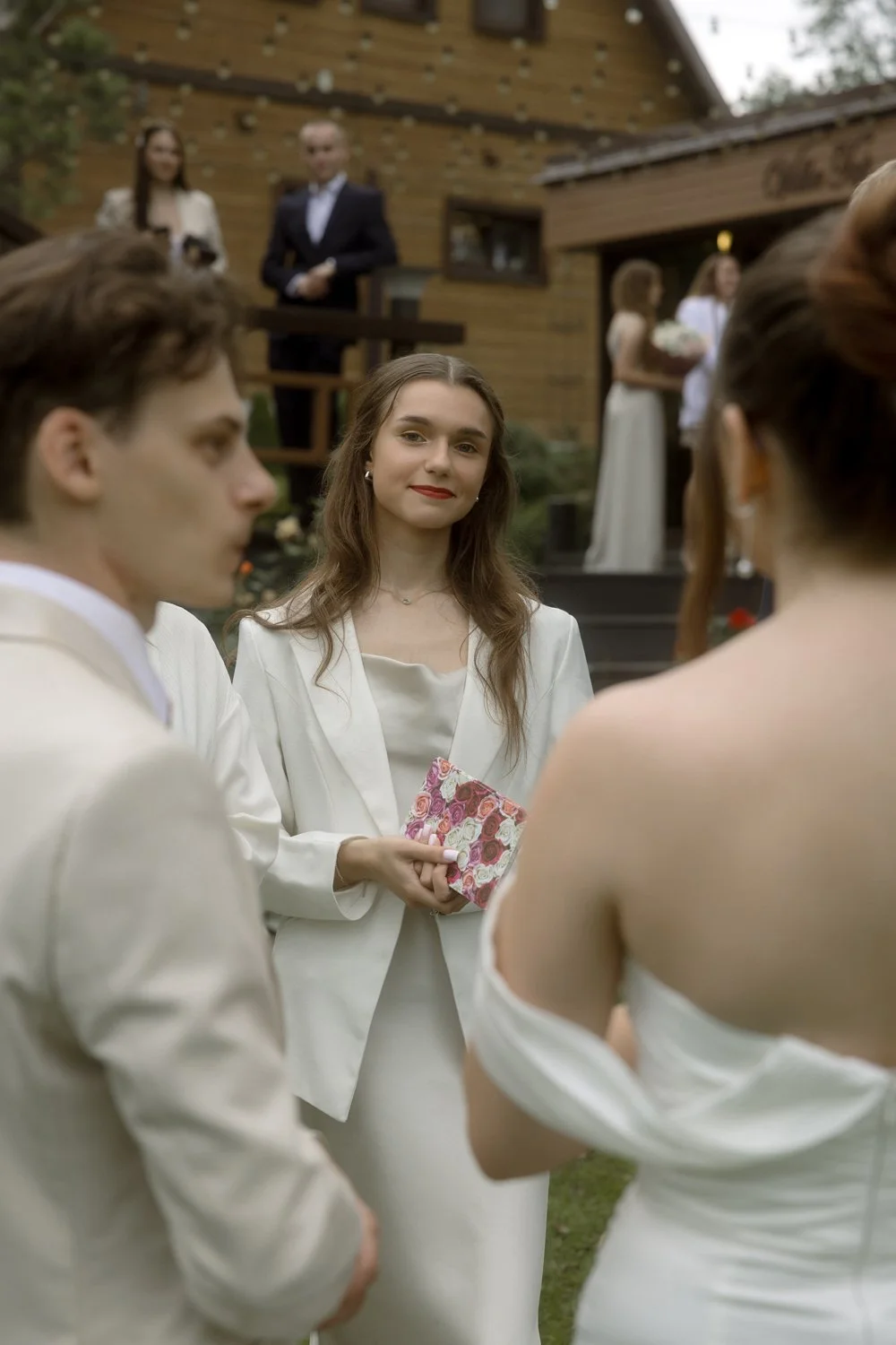 A woman in a white blazer holding a floral-patterned gift box, standing between a man and a woman in white dresses, at an outdoor wedding or similar ceremony, with people and a wooden building in the background.