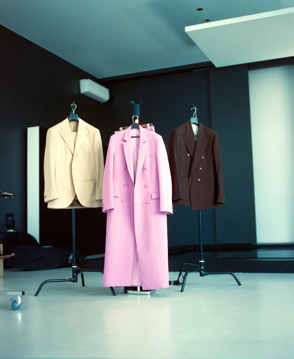 Three mannequins wearing blazers in cream, pink, and brown are displayed on stands in a room with dark walls and minimal lighting.