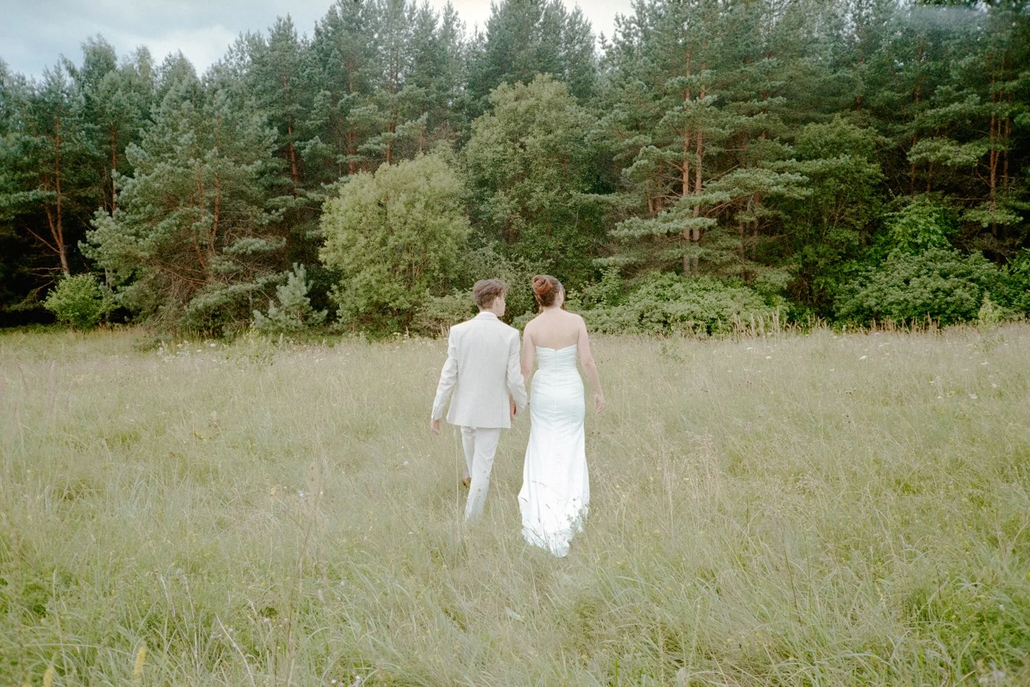 A couple in wedding attire walking through a grassy field towards a forested area, seen from behind.