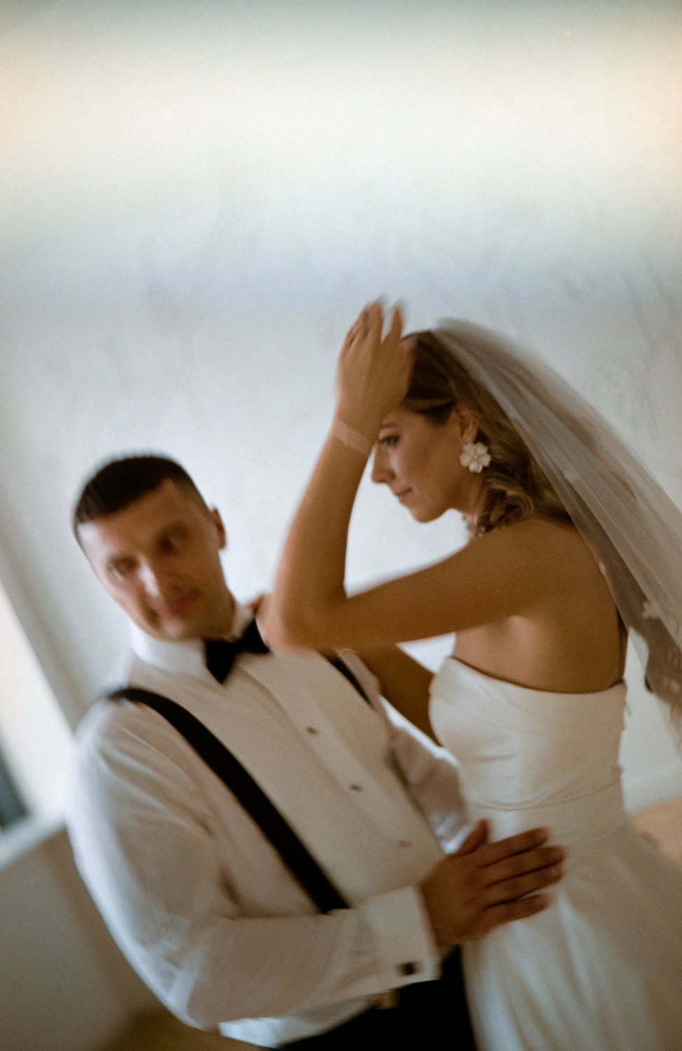 Blurred photo of a bride in a white wedding dress and veil and a groom in a tuxedo with suspenders, standing close together in an indoor setting.