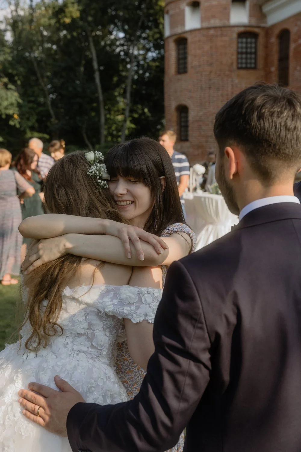 A woman in a wedding dress happily hugging a young girl dressed in a floral dress, while a man in a suit looks on at an outdoor wedding celebration. In the background, there are people mingling near a brick building.