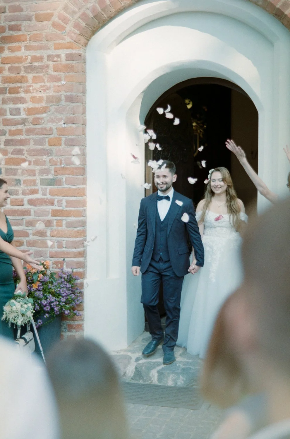 Bride and groom walking out of a church as guests throw flower petals, celebrating a wedding.