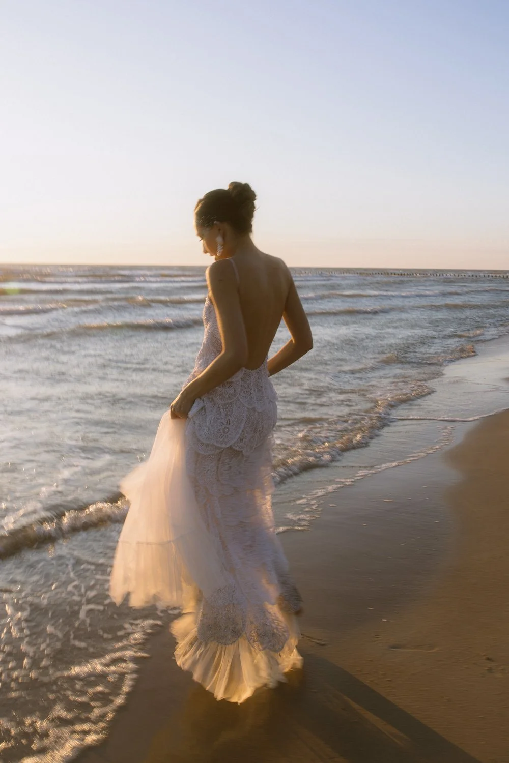 A woman wearing a backless white lace dress walking along the beach at sunset.