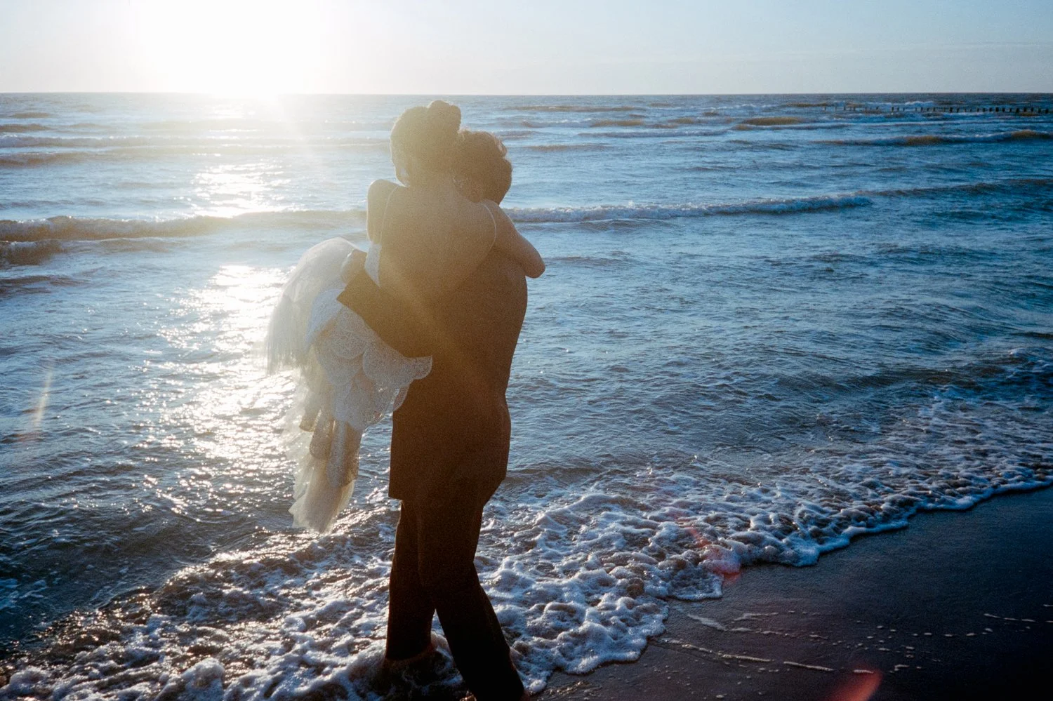 A person carrying a child on their back along the shoreline at the beach during sunset.
