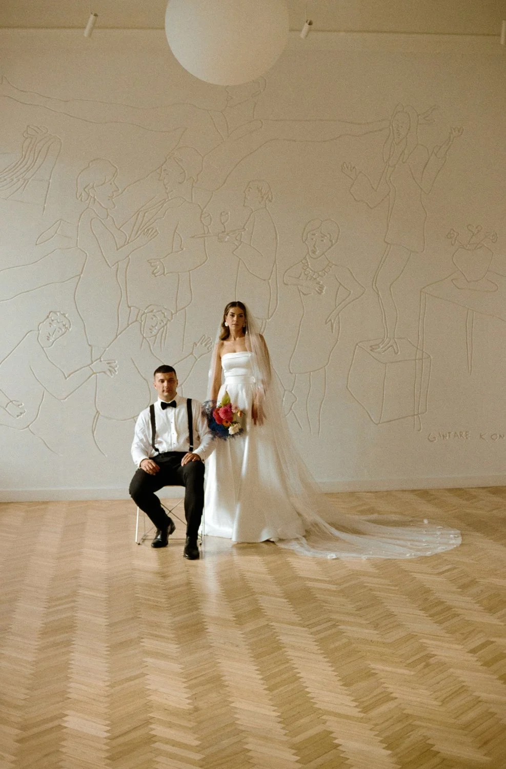 A bride and groom pose in a spacious room with a wooden floor and a large wall mural. The bride is standing in a white wedding dress holding a bouquet, and the groom is seated on a chair in a white shirt with suspenders and a bowtie.