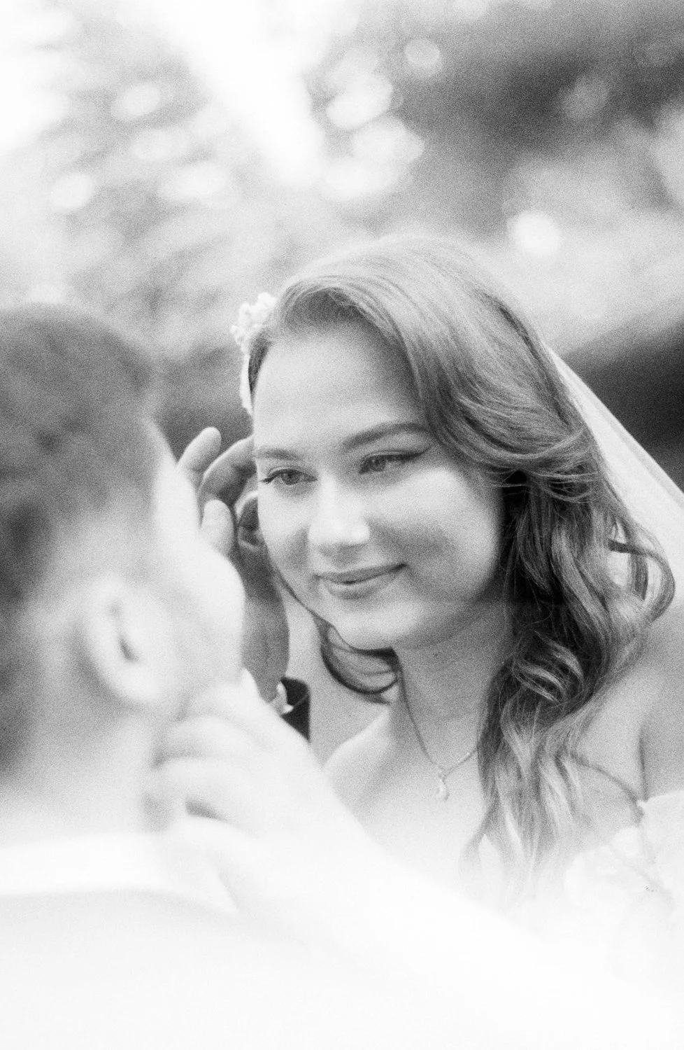 A black-and-white photo of a woman with long, wavy hair smiling gently as she looks at a man, with a blurred background of bokeh lights.