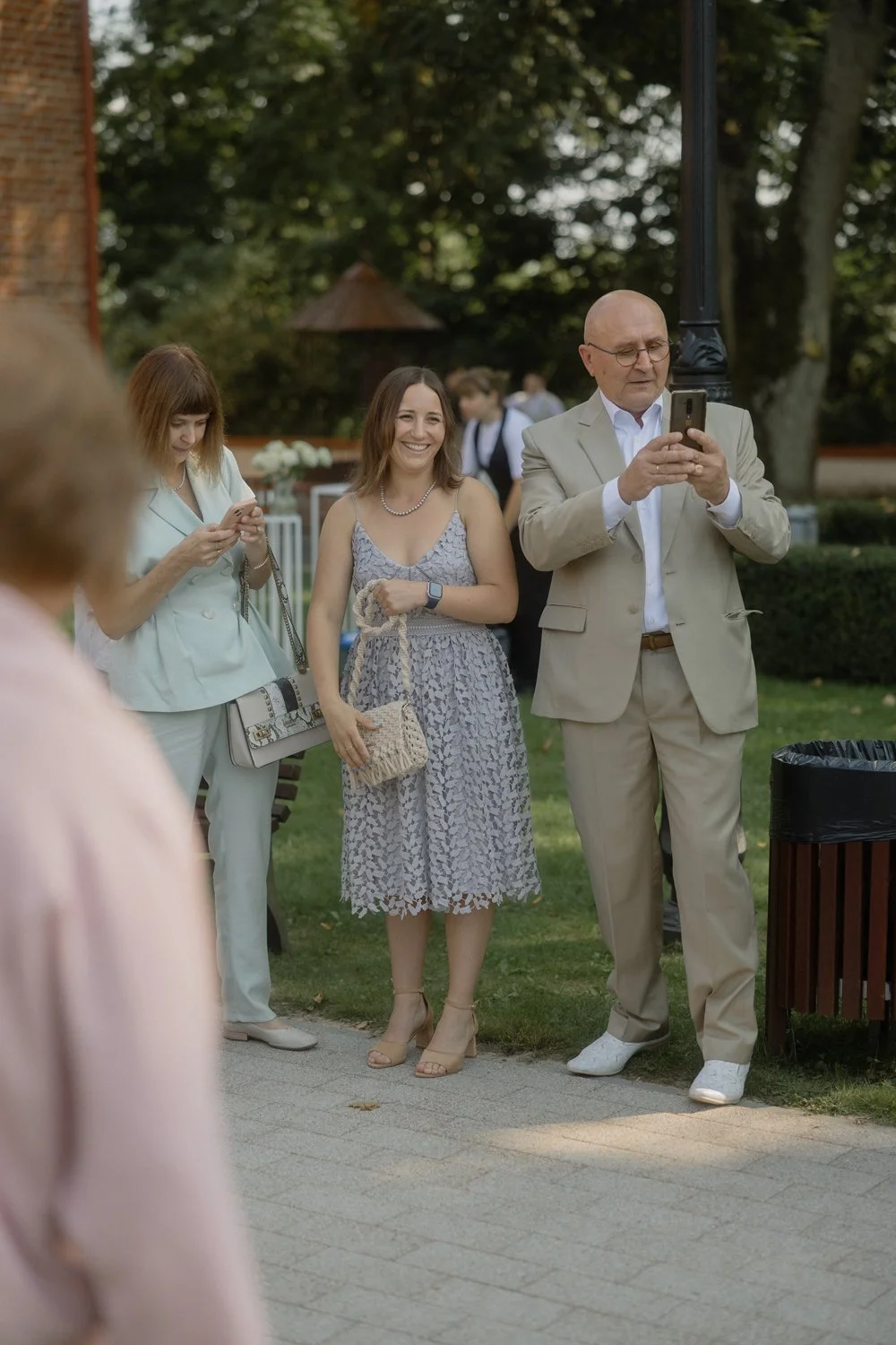 People dressed in formal attire at an outdoor event, with a man in a beige suit taking a photo with his phone, and women smiling and using their phones.