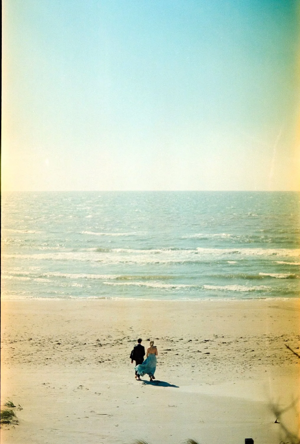 A couple walking on a beach toward the ocean, with the man dressed in a dark suit and the woman in a flowing blue dress, under a clear sky.