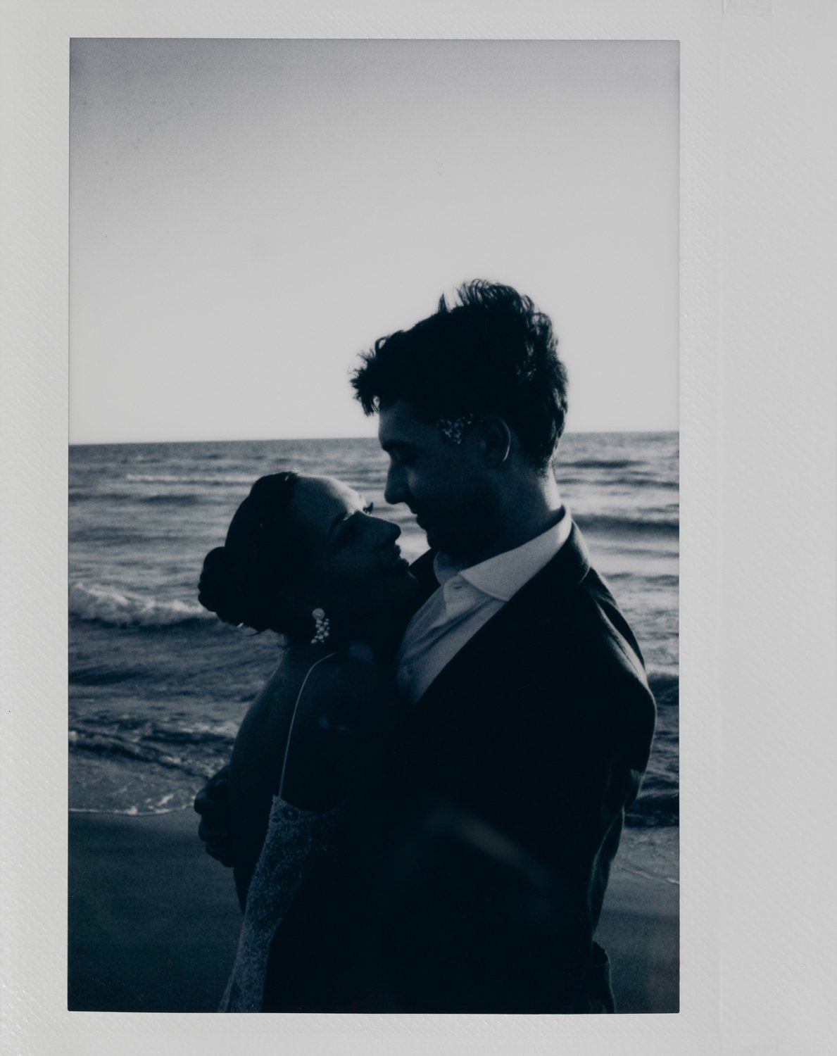 A black and white photo of a couple embracing by the ocean, looking into each other's eyes.