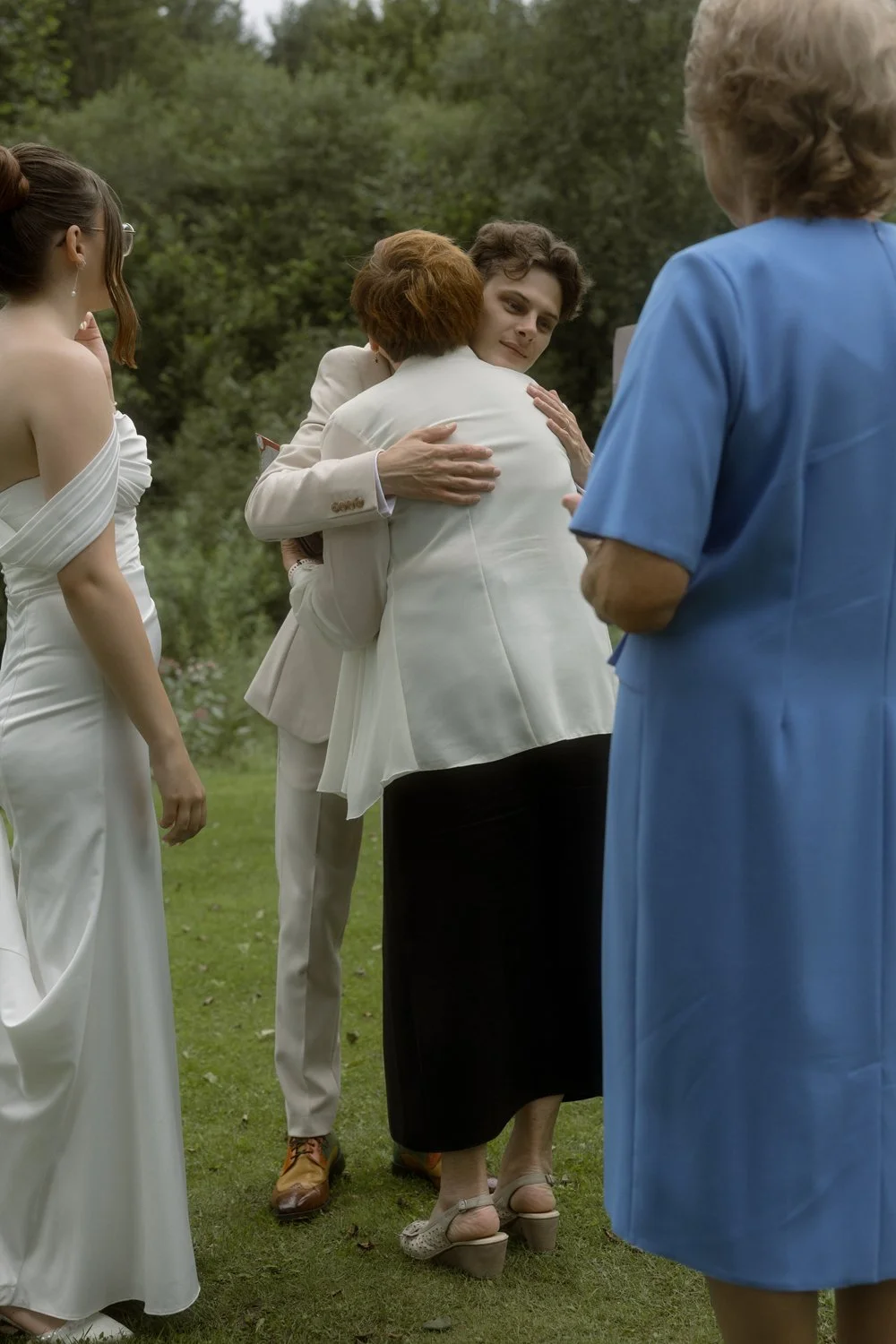 Two women hugging, one wearing a white blazer and black skirt, the other in a white dress, an outdoor gathering with three other women nearby.