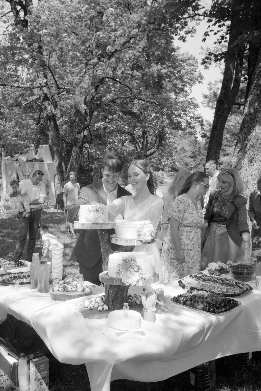 People gathered outdoors at a celebration, with a couple cutting a wedding cake on a table filled with desserts and drinks, under trees in the background.