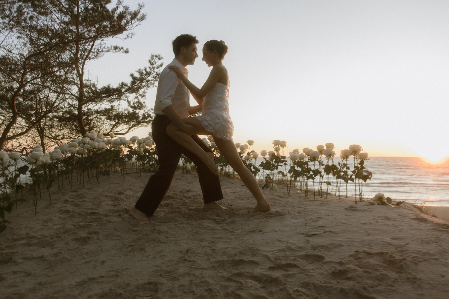 A couple dancing on the beach at sunset, with the man holding the woman in his arms and the woman leaning towards him. The scene features white flowers and trees in the background.