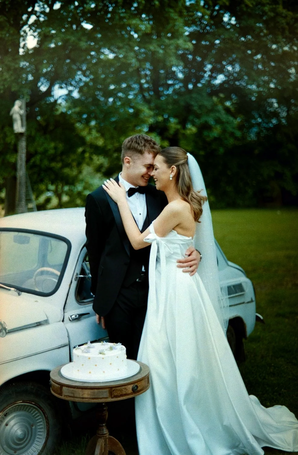 A bride and groom in wedding attire embracing outdoors next to a vintage car with a wedding cake on a small wooden table in front of them.