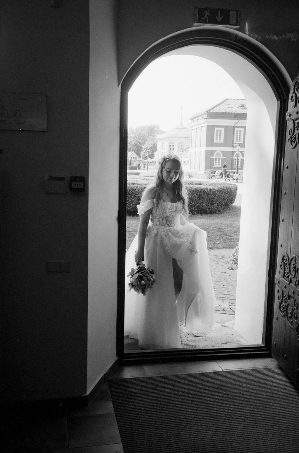 A bride stands on the doorway outside, holding a bouquet, as she looks down and smiles. She wears a wedding dress with off-the-shoulder sleeves, and the scene is captured in black and white.