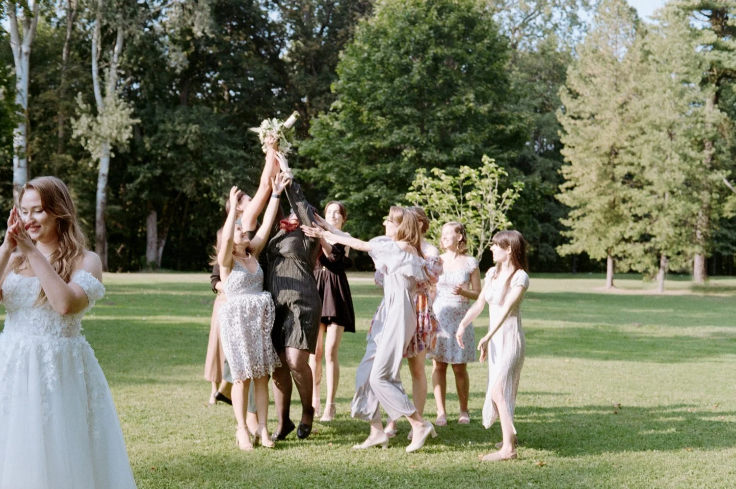 A group of women celebrating outdoors in a park with trees, some women are holding a bouquet while others are clapping and smiling.