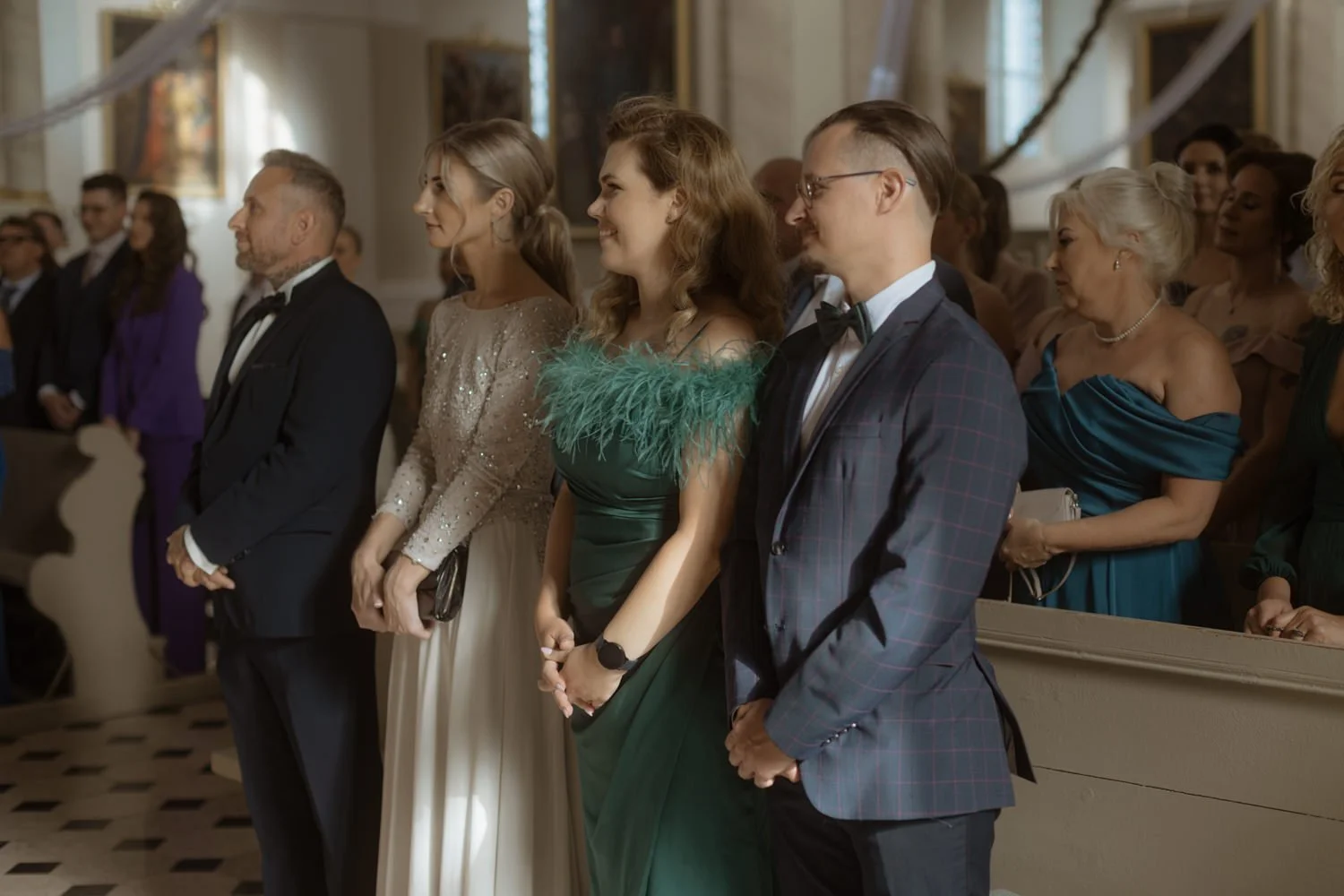 People standing in line at a formal event inside a decorated church, dressed in elegant attire including tuxedos and evening gowns, some smiling and looking forward.