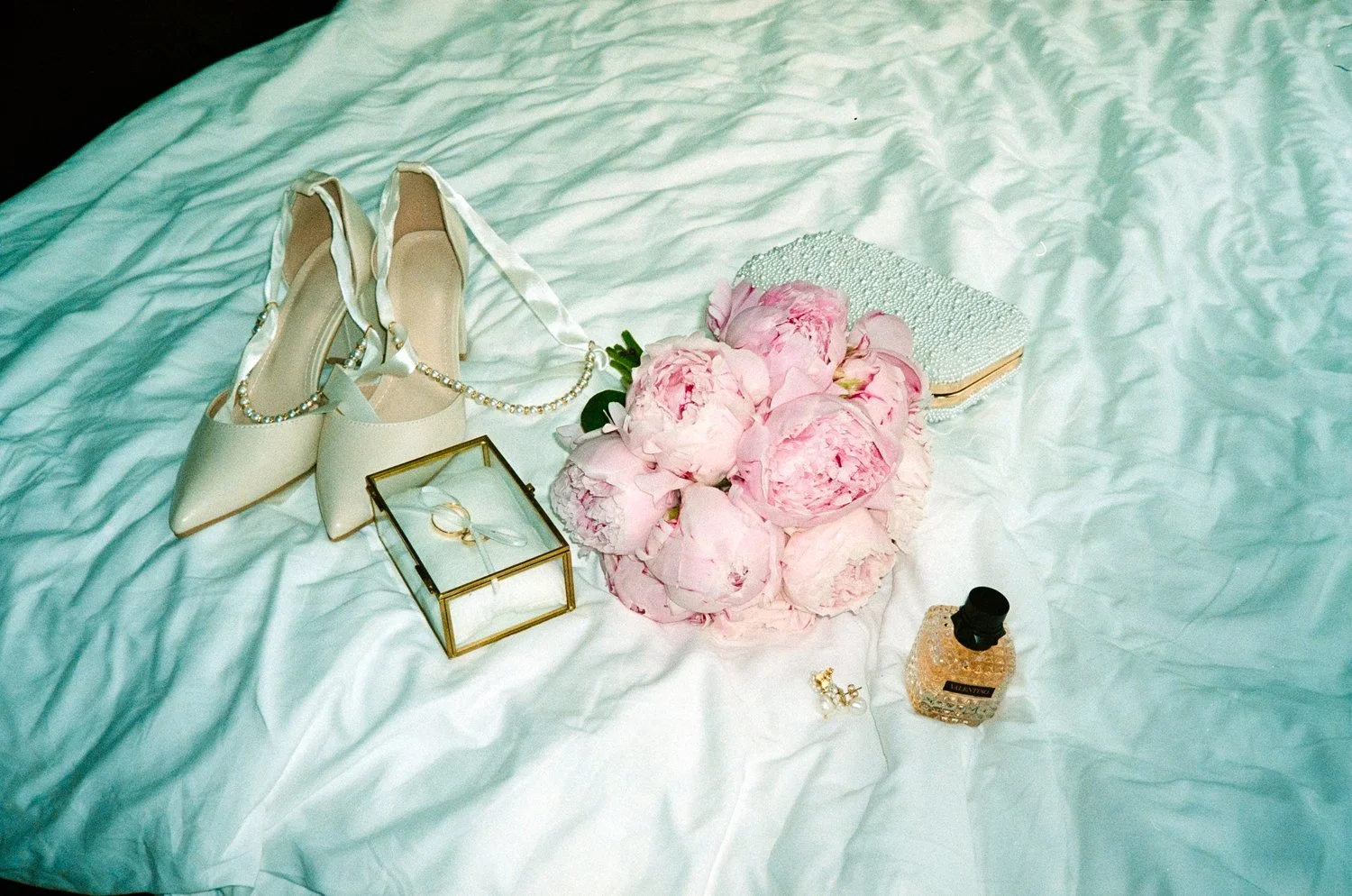 A collection of wedding accessories on a white bedsheet, including a pair of white high-heeled shoes, a pearl necklace, a small glass jewelry box with rings inside, a bouquet of pink peonies, a pearl-embellished clutch, a small perfume bottle, and a 