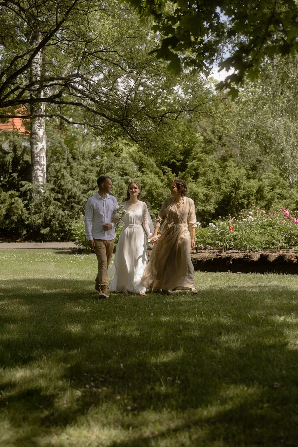 Three people walking through a lush green park on a sunny day, with trees and flowers surrounding them, holding hands and smiling.