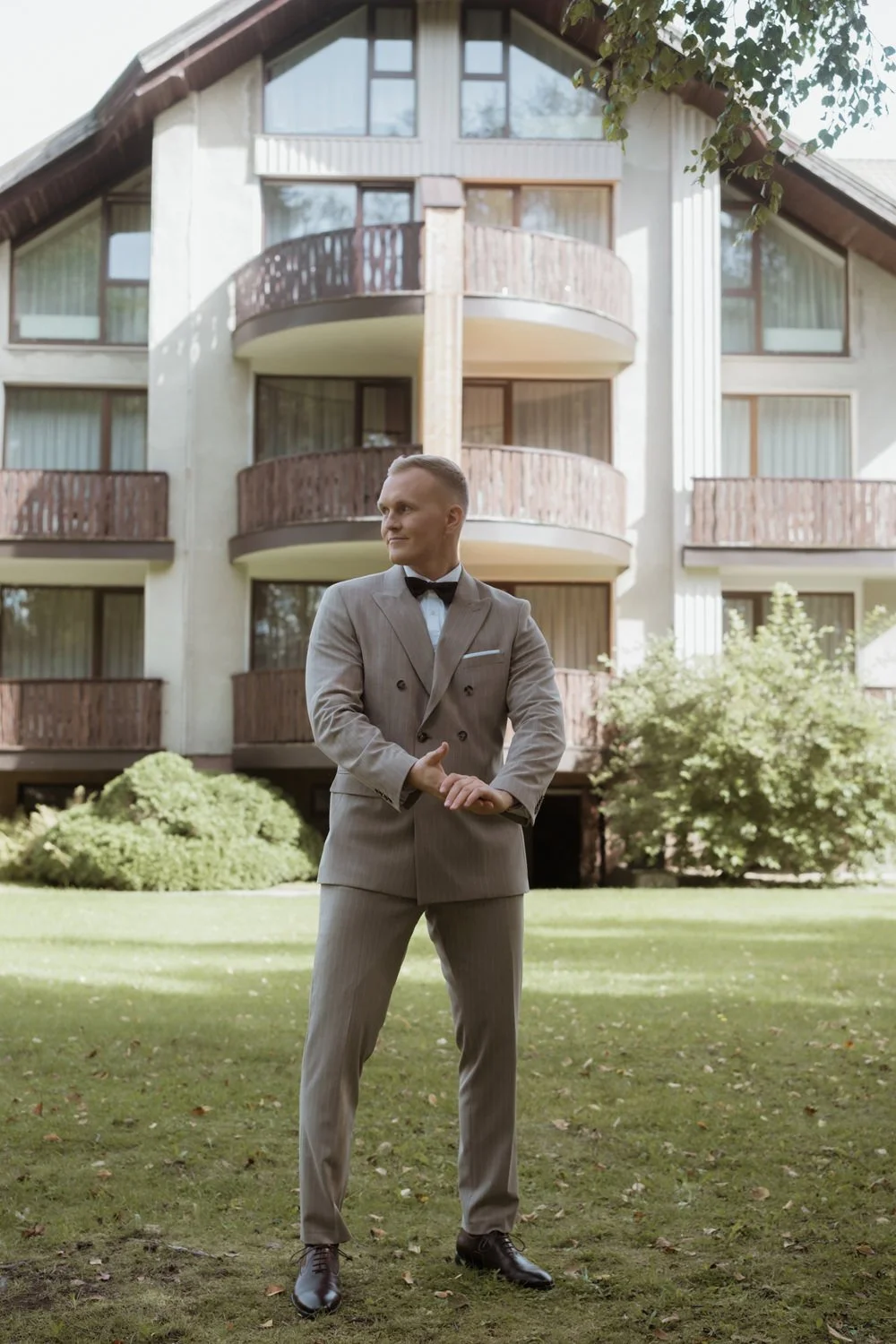 Man in a gray suit with a black bow tie standing outdoors on a grassy lawn in front of a modern house with balconies and large windows.