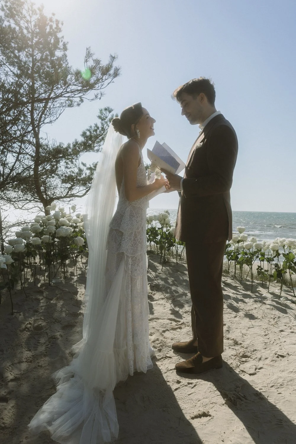 A bride and groom stand on a sandy beach, holding hands during their wedding ceremony, with the ocean and a clear sky in the background.