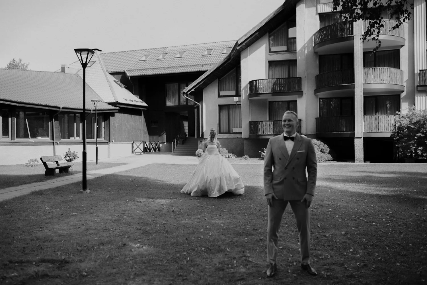 Black and white photo of a bride and groom outside a building with balconies. The groom is in the foreground wearing a suit with a bow tie, smiling. The bride is in the background, wearing a wedding dress, holding a bouquet, and smiling.