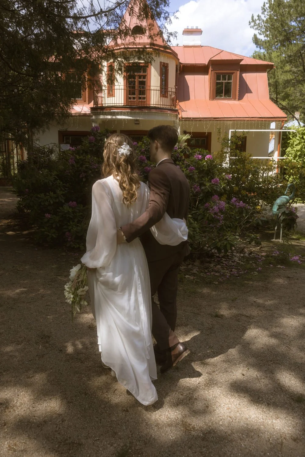 Couple in wedding attire walking hand in hand in a garden with a pink house in the background.