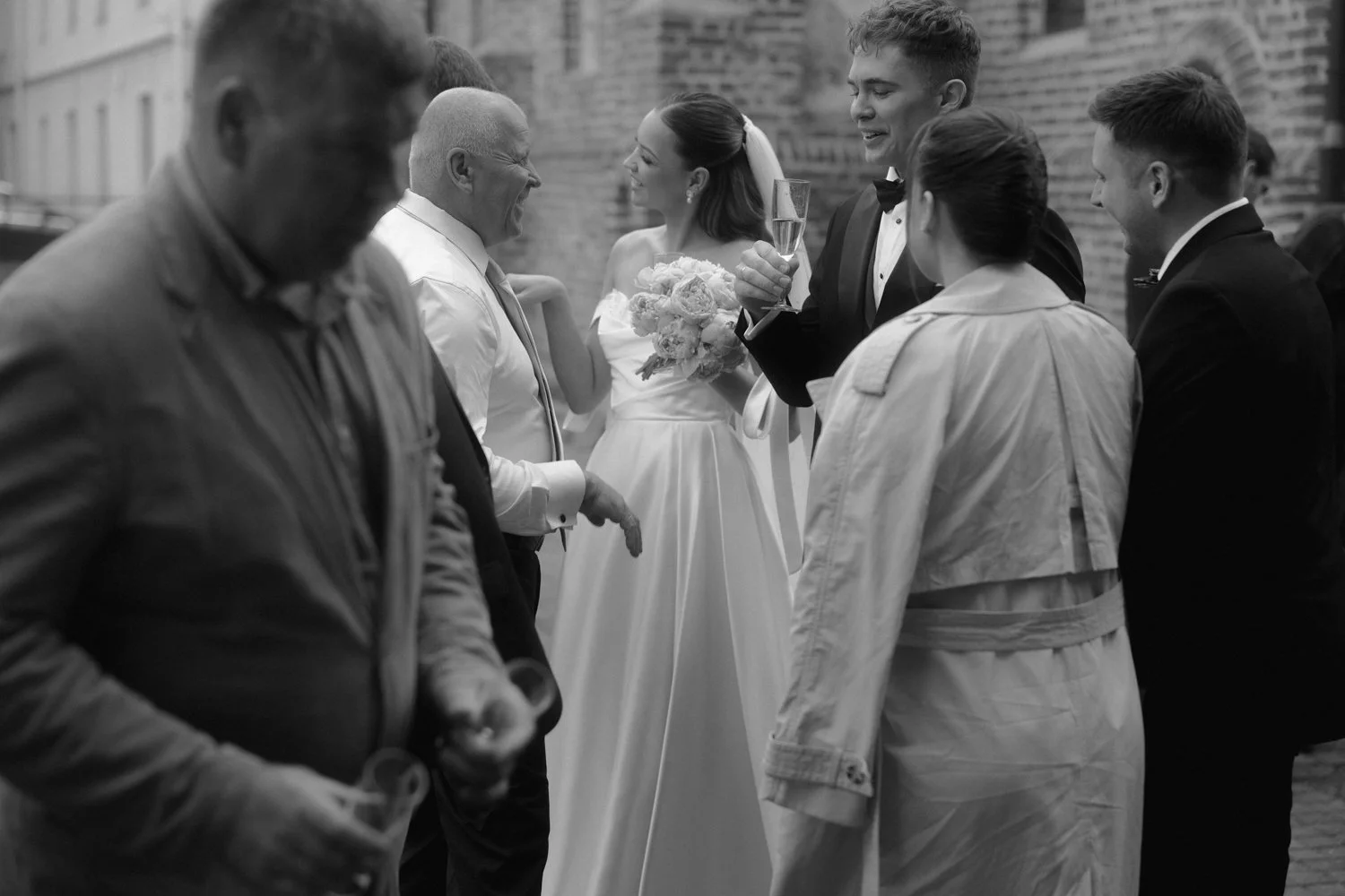 A wedding reception with a bride holding a bouquet and dressed in a white gown, surrounded by guests including men in suits and a woman in a trench coat, in an indoor space with brick walls.