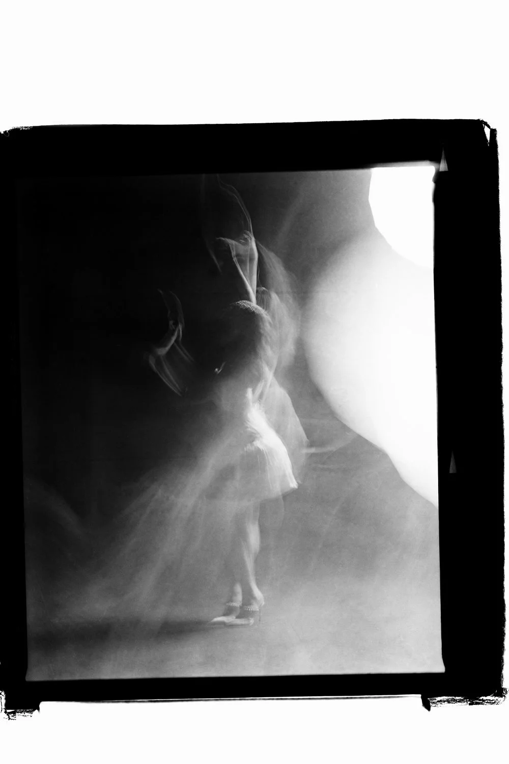 Black and white photo of a person with long hair, captured in a moment of movement, with light illuminating their face and upper body, against a dark background.