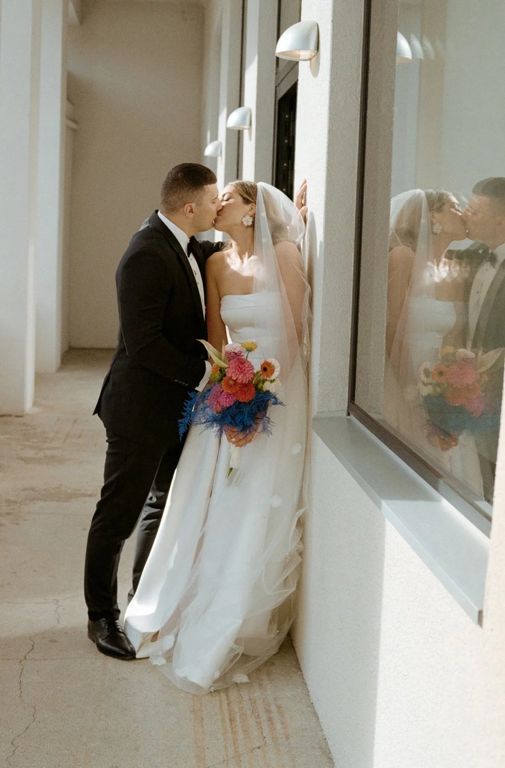 A bride and groom share a kiss while standing beside a large window. The bride is holding a colorful bouquet and wearing a strapless white wedding gown with a veil. The groom is dressed in a black tuxedo with a bow tie. The reflection of the couple c