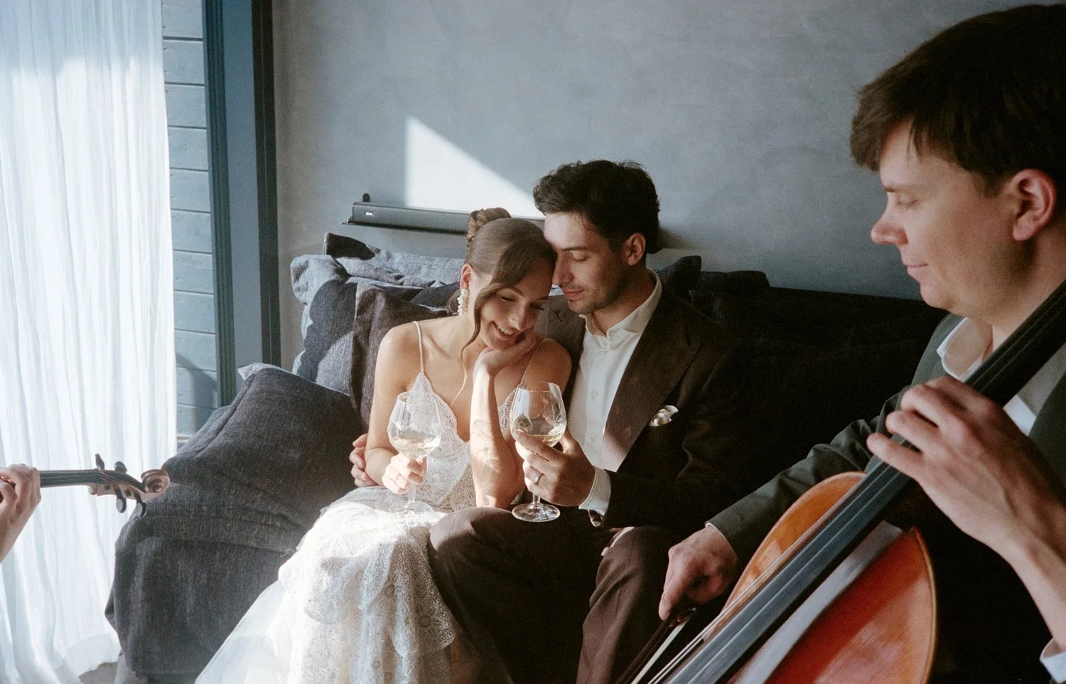 A bride and groom sharing a moment with a musician and a cellist, holding wine glasses indoors near a window.