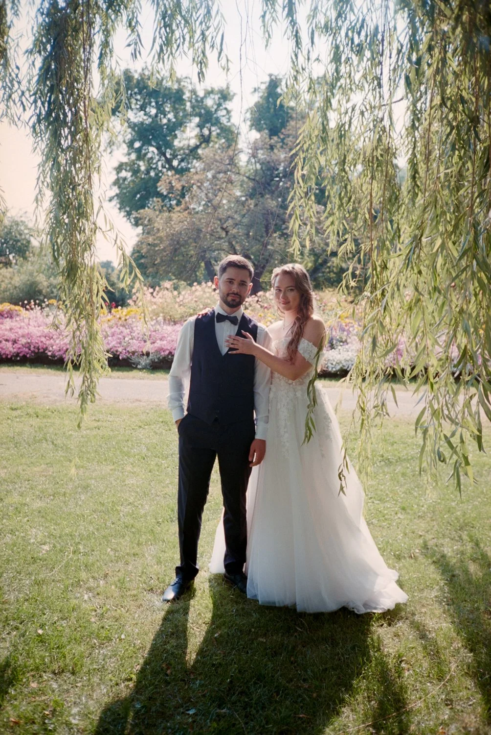 A bride and groom standing under a tree branch in a park with colorful flowers in the background, dressed in wedding attire.