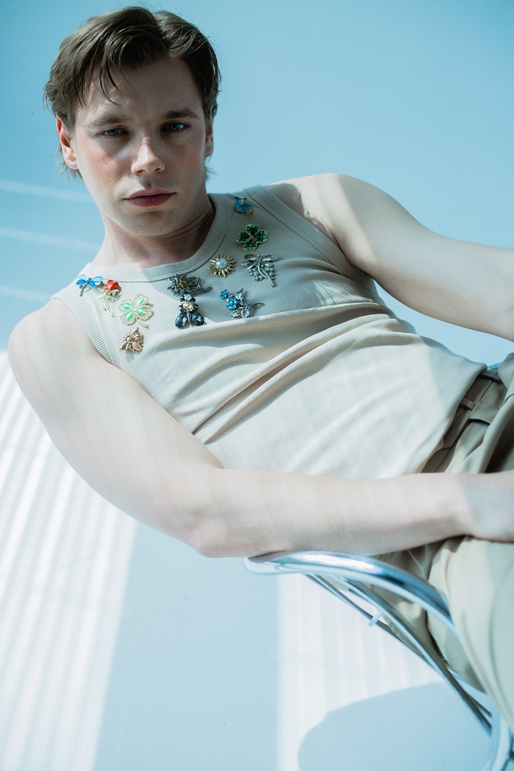 A young man with light brown hair and blue eyes wearing a beige tank top decorated with colorful floral and clover-shaped pins. He is sitting outdoors against a bright blue sky.