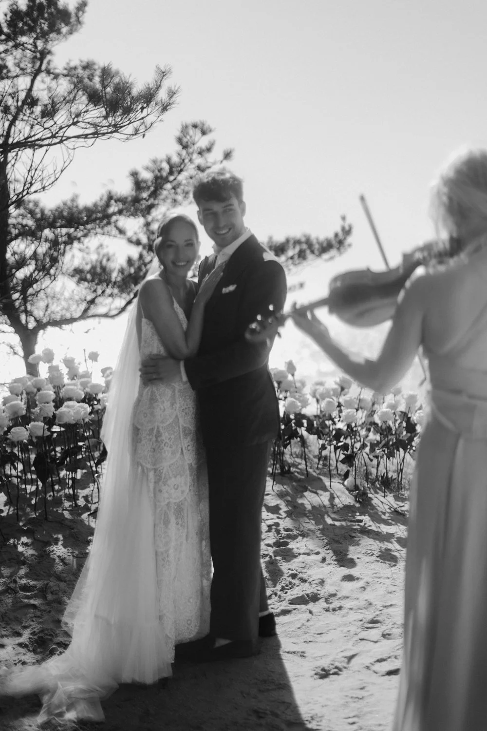 A wedding couple standing in a field of flowers, with a musician playing the violin nearby, on a sunny day.