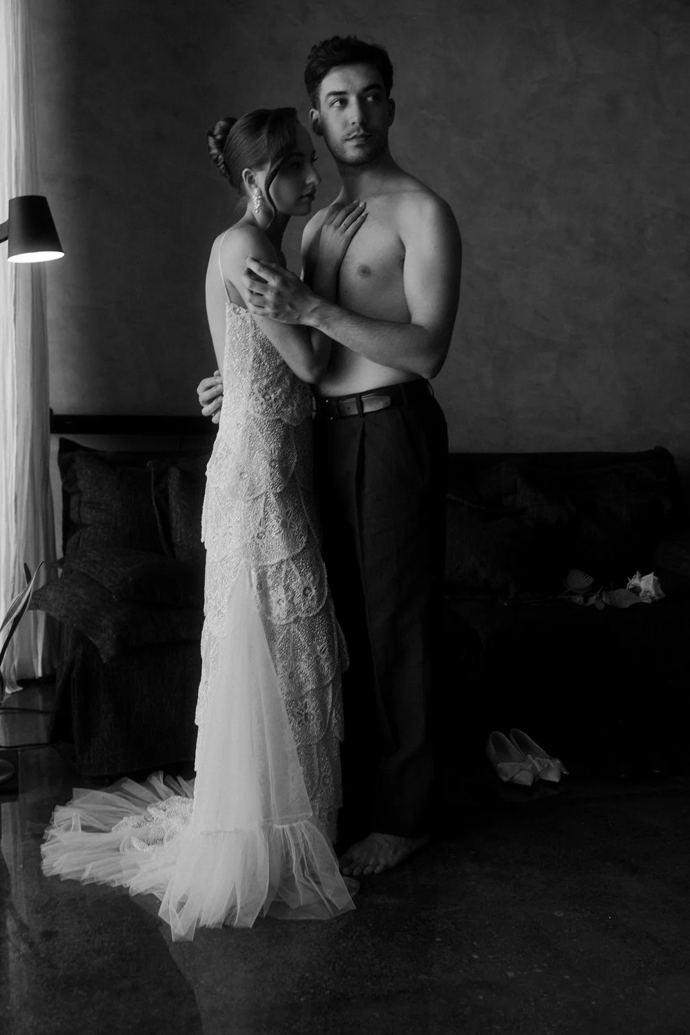 A black-and-white photo of a young couple in an intimate embrace, the woman in a wedding dress and the man shirtless, standing close in a dimly lit room.