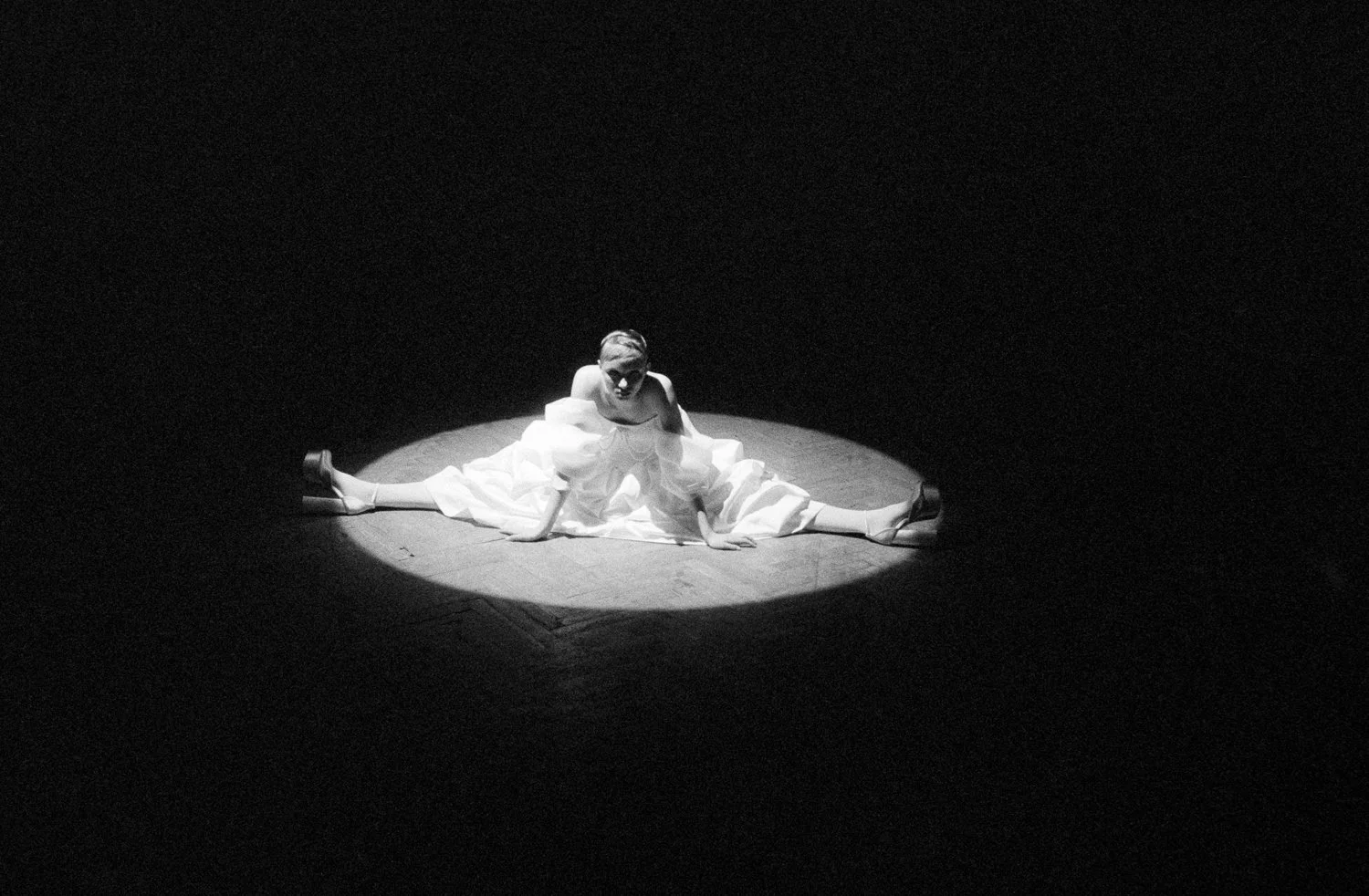 A woman performing on stage in a dramatic pose, sitting with her legs spread wide and leaning forward, illuminated by a spotlight in a dark theater.