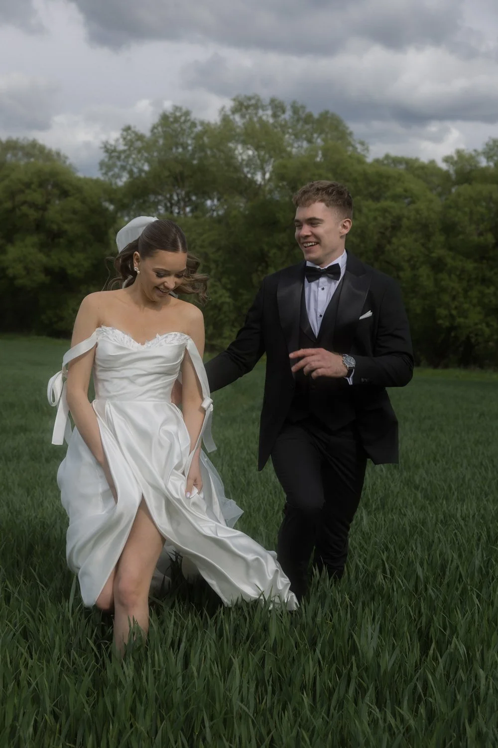 A bride and groom laughing and walking together in a green field outdoors, with trees and a cloudy sky in the background. The bride wears a white off-the-shoulder wedding dress, and the groom wears a black tuxedo with a bow tie.