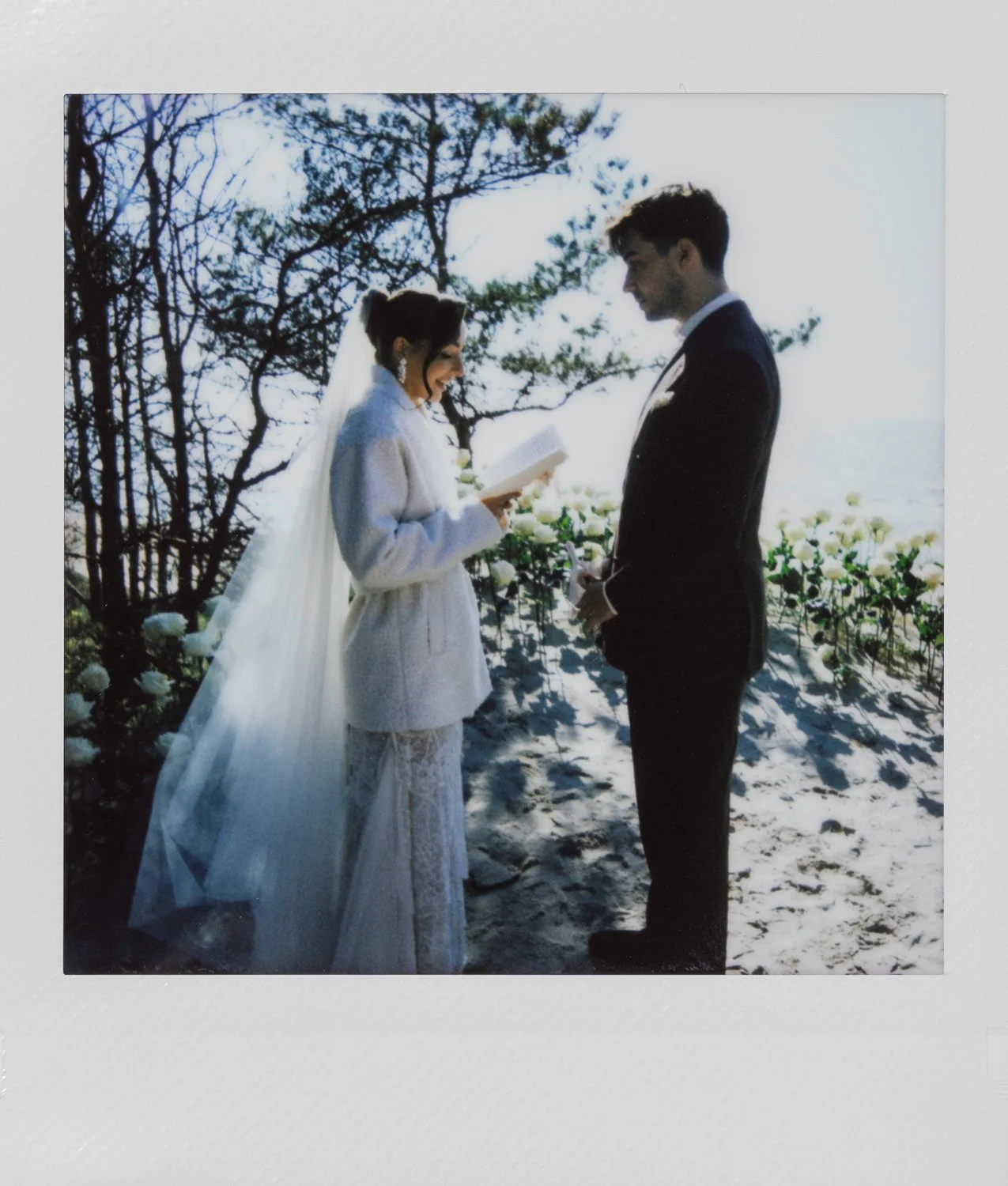 A bride and groom exchanging vows outdoors, with the bride reading from a piece of paper, surrounded by trees and white flowers.